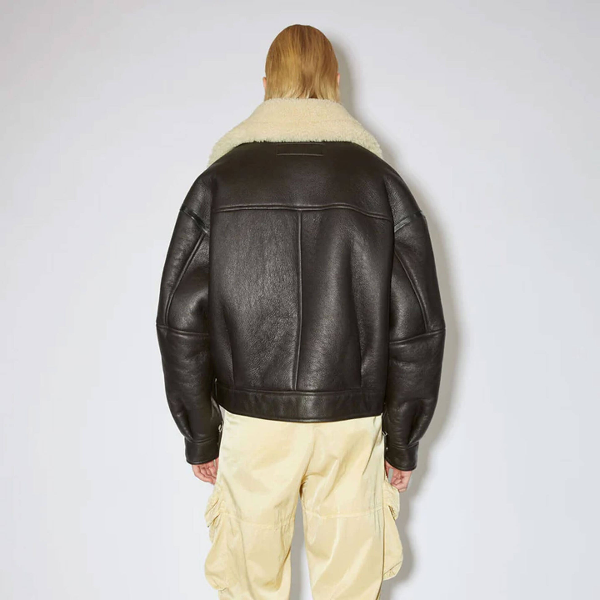 Back view of a women’s leather shearling aviator coat, crafted from black cowhide leather with faux shearling lining and oversized shearling collar. The design showcases a belted waist, structured shoulders, and a relaxed aviator cut, displayed on a female model to emphasize durability, fashion appeal, and comfort for winter streetwear outfits.